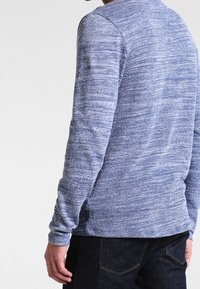 Chemise à manches longues bleu chiné avec un motif tricoté texturé. Tissu lisse, encolure ronde et coupe ajustée. Portée avec un jean foncé.