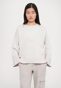 Lys beige oversized sweater med brede ærmer, der har en dyb halsudskæring. Lavet af blødt stof med ribbede manchetter og kant.
