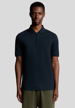 Junger Mann trägt ein marineblaues Polohemd mit kurzen Ärmeln und Reißverschluss sowie olivgrüne Hosen, steht vor einem einfarbigen grauen Hintergrund.