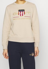 Sweat-shirt beige à col rond, avec un logo écusson rouge et marine et une broderie "GANT". Tissu texturé avec poignets et ourlet côtelés.