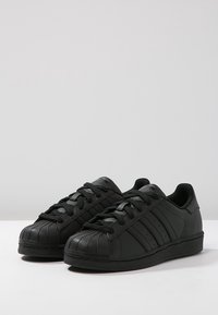 Černé sportovní tenisky s lesklým koženým povrchem, gumovou špičkou a třemi postranními pruhy. Mají ploché tkaničky a strukturovanou podešev.