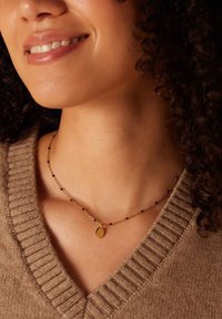 Collier en or avec de petites perles noires et un pendentif ovale, reposant sur un pull en maille léger marron clair. Gros plan sur la zone du cou.