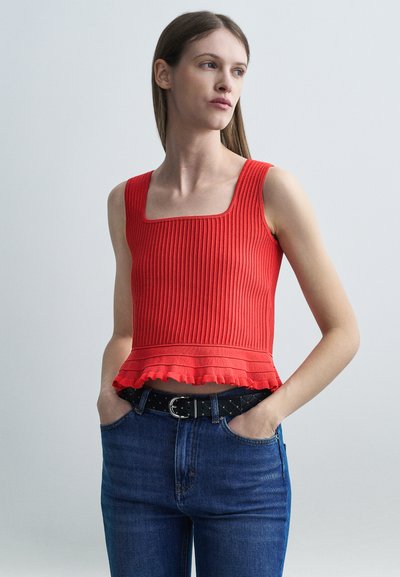 Jeune femme portant un haut rouge sans manches à côtes avec un ourlet volanté et un jean bleu, les mains dans les poches, regardant sur le côté contre un fond uni.