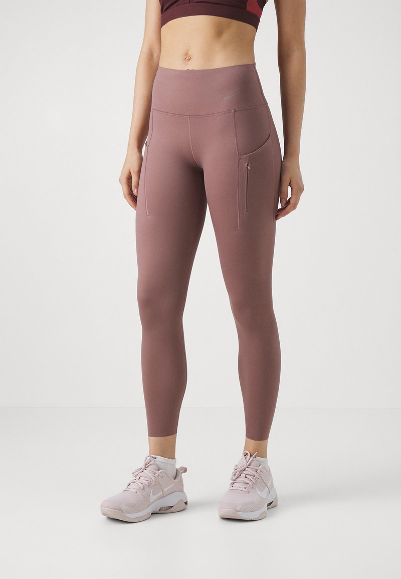 Leggings de talle alto, ajustados, en color mauve, con bolsillos laterales con cremallera, textura suave y detalles cosidos, combinados con zapatillas deportivas de color claro.