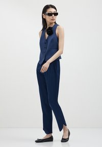 Femme portant un gilet sans manches bleu foncé avec une épingle à fleurs noires, un pantalon assorti avec des fentes sur les côtés, des chaussures plates noires et des lunettes de soleil rectangulaires.