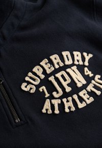 Sudadera con cremallera de color azul marino con texto bordado en textura crema que dice "SUPERDAY 7 JPN 4 ATHLETIC." Presenta una cremallera de metal.