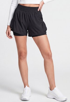Person iført sorte lagdelte træningsshorts med elastisk talje og hvide sneakers, stående med den ene hånd på hoften.