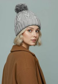 Gray knitted beanie with cable pattern and pom-pom on top. Model wears a brown coat, showcasing the hat's texture and design.