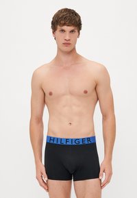 Schwarze Boxershorts mit einem blauen Bund, der das "HILFIGER"-Logo trägt. Glattes Material, enganliegendes Design und Länge bis zur Mitte des Oberschenkels.