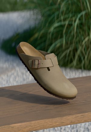 Beige sabbi con finitura opaca, con una fibbia regolabile marrone e suola testurizzata, esposti su una superficie in legno con verde sullo sfondo.