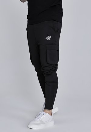 Jogging noir avec poches cargo, design fuselé et détail logo blanc sur la hanche. Associés à des baskets blanches. Texture lisse.