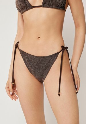 Kvinde iført en brun metallisk bikini med justerbare sidelukninger med guldfarvede perler på en lys baggrund.