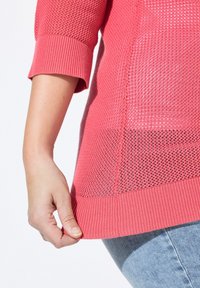 Top de punto en color coral claro con un diseño de malla texturizada y puños acanalados. Dobladillo ligeramente recogido, revelando un efecto de capas. Material suave y ligero.