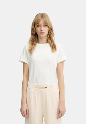 Donna bionda con capelli sciolti che indossa una camicia bianca a maniche corte e pantaloni beige a vita alta con cintura con fibbia dorata, in posizione neutra.