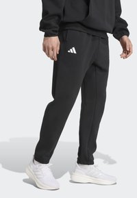 Crne sportske hlače uskog kroja, s bijelim Adidas logotipom na lijevom bedru i bočnim šavovima. Izrađene od mekog materijala.