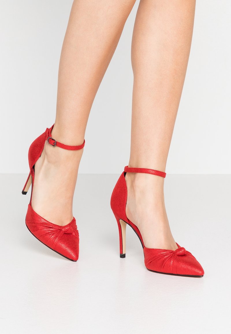 Jambes portant des escarpins rouges à bout pointu avec des brides à la cheville et un détail nœud sur un fond clair uni.