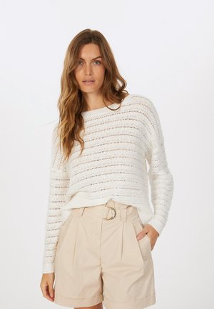 Weißer gestrickter Pullover mit lockerem Schnitt und horizontalen Streifen, kombiniert mit hoch taillierten beigen Shorts, die einen Gürtel mit einer runden Schnalle haben.