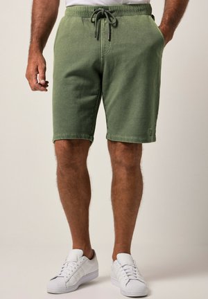 Mand iført olivengrønne shorts med snøre og hvide sneakers, stående med den ene hånd i lommen foran en ensfarvet baggrund.