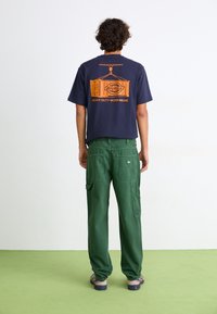Navy cotton t-shirt featuring an orange graphic print on the back. Paired with green cargo pants with multiple pockets and a relaxed fit.