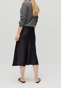 Musta satiini midi-hame, jossa on virtaava siluetti ja sileä rakenne, yhdistettynä harmaaseen villaneuleeseen ja mustiin liukuhihnasandaaleihin.