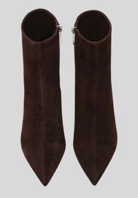 Un paio di stivaletti in suede marrone scuro con punta affusolata e cerniere interne argentate, esposti in posizione verticale su uno sfondo grigio chiaro.