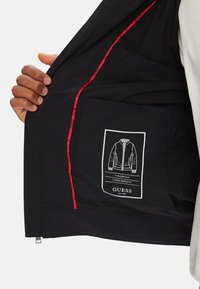 Interno della giacca nera con accenti rossi, dotato di una tasca e di un'etichetta che dettaglia le proprietà antivento e idrorepellenti.