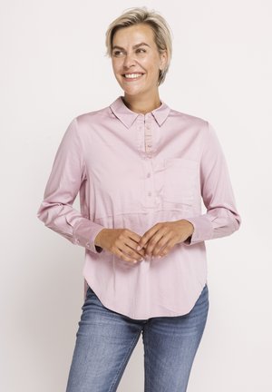 Blonde Frau lächelnd, trägt ein hellrosa langärmliges Hemd mit Knöpfen und Kragen sowie blaue Jeans, steht vor einem weißen Hintergrund.