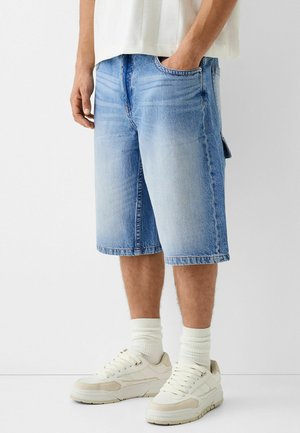 Hombre vistiendo pantalones cortos de mezclilla azul claro hasta la rodilla, calcetines blancos y zapatillas blancas, de pie contra un fondo blanco liso.