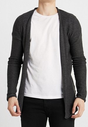 Homme portant un t-shirt blanc sous un cardigan en tricot gris foncé ouvert, associé à un pantalon noir, debout devant un fond uni.
