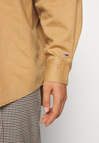 Camicia khaki con bottoni dal tessuto liscio, con un patch con logo sulla manica. Pantaloni a motivo houndstooth parzialmente visibili.