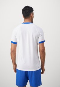 Hvid kortærmet atletisk skjorte med blå halsudskæring og ærmeaccent, der har et tekstureret mønster. Båret over blå atletiske shorts.