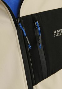 Beige and black jacket featuring blue zipper pulls, with two zippered pockets on the front and contrasting fabric textures.