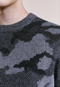 Grå stickad tröja med ett texturerat mönster i mörkare gr å nyanser. Rund halsringning och ribbad mudd, vilket framhäver en mjuk ullblandning.