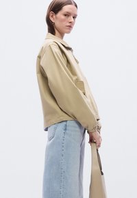 Ανοιχτό μπεζ μπουφάν με γιακά, κλείσιμο με φερμουάρ και πλαϊνές τσέπες. Φορεμένο πάνω από μπλε τζιν παντελόνια, κρατώντας μια μπεζ τσάντα. Μινιμαλιστικός σχεδιασμός.