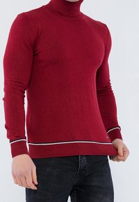 Czerwony sweter typu golf z dopasowanym krojem, prążkowaną fakturą oraz kontrastującymi białymi i czarnymi paskami na dole i mankietach.