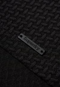 Tissu noir texturé avec un motif tissé, agrémenté d'une étiquette noire mate portant l'inscription "Superdry".