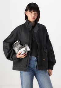 Čierna oversized denimová bunda s vysokým golierom, detailmi na gombíky a rebrovanými manžetami, v kombinácii s textúrovanou striebornou kabelkou a modrými džínami.