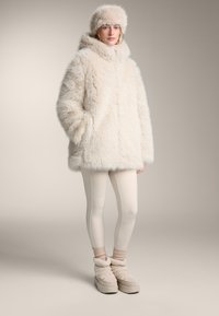 Krémový kabát z umelého kožušina s vysokým golierom, klobúk z toho istého materiálu, béžové legíny a plyšové nazúvacie topánky. Jemný, štruktúrovaný materiál po celom povrchu.