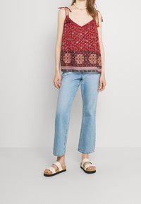 Top in tessuto floreale rosso con scollatura a V e spalline regolabili, abbinato a jeans svasati di colore blu chiaro e sandali crema con dettagli in sughero.
