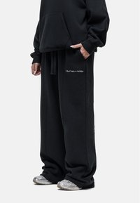 Zwarte sweatshirt met een voorzak en zwarte sweatpants met een wit geborduurd logo. Materiaal lijkt zacht en textuur heeft een fijne structuur, pasvorm is los.