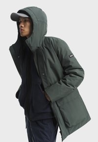 Zelená nepremokavá bunda s kapucňou, so zipsom a patentkami, bočnými vreckami a nášivkou s logom na rukáve. Nosená cez námornícku fleecovú mikinu.