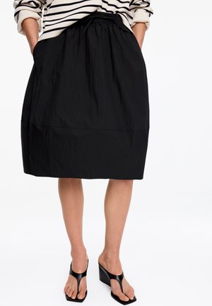 Žena má na sebe čiernu sukňu po kolená s predným sťahovaním na šnúrku, kombinovanú s čiernymi sandálmi na klinovom opätku a pruhovaným dlhým tričkom s vyhrnutými rukávmi.