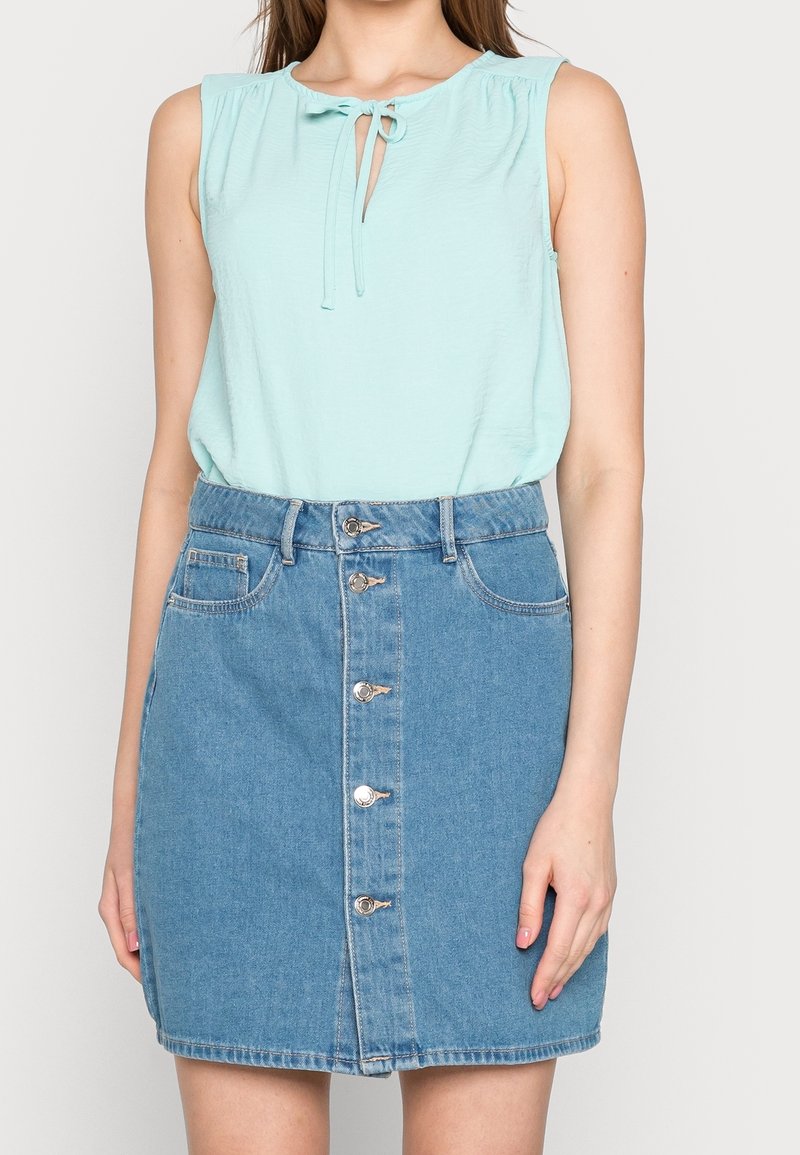 Top senza maniche di un azzurro chiaro con un nodo al collo, abbinato a una gonna di denim con bottoni frontali e tasche, in un lavaggio azzurro chiaro.