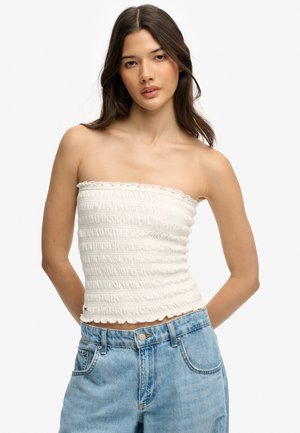 Jeune femme aux longs cheveux foncés, portant un haut blanc texturé sans bretelles et un jean bleu en denim, debout devant un fond blanc.