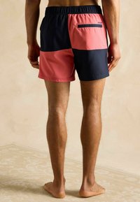 Mężczyzna w kąpielówkach w kolorze koralowo-różowym i granatowym, stoi boso na wzorzystej beżowej podłodze.