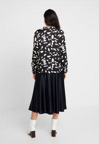 Blouse noire avec des motifs abstraits blancs, manches longues et ourlet arrondi, associée à une jupe en satin bleu marine fluide et des bottines blanches.