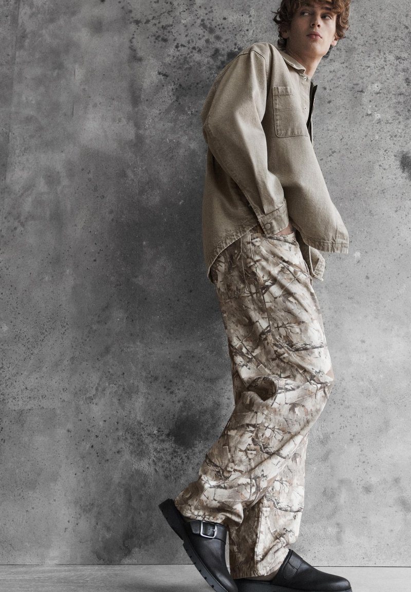 Jeune homme portant une chemise beige, un pantalon cargo camouflage et des chaussures noires à boucles, debout devant un mur en béton gris texturé.