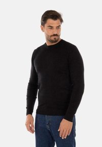 Maglione nero a maglia con scollatura rotonda e maniche lunghe, caratterizzato da un orlo a coste. Indossato con jeans di denim blu, che presentano una vestibilità aderente.