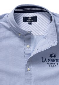 Vaaleansininen nappipaita, jossa on muotoiltu kaulus, kirjailtu logo ja kontrastivärinen raidallinen sisäkaulus. Paidassa on valkoiset napit ja yksityiskohtaiset ompeleet.