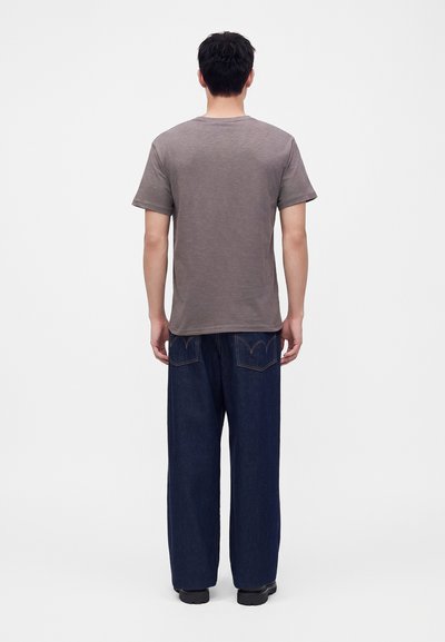 Homme portant un t-shirt marron uni à manches courtes et un jean bleu foncé à jambes larges, debout de dos face à la caméra sur fond blanc.